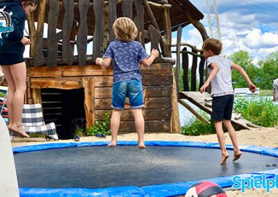 Zwei Kinder springen auf dem Trampolin, was gleich neben dem Holzspielplatz steht