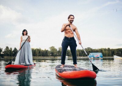 Muskulöser Mann und Frau im Brautkleid fahren auf einem Stand-Up-Paddle auf dem See
