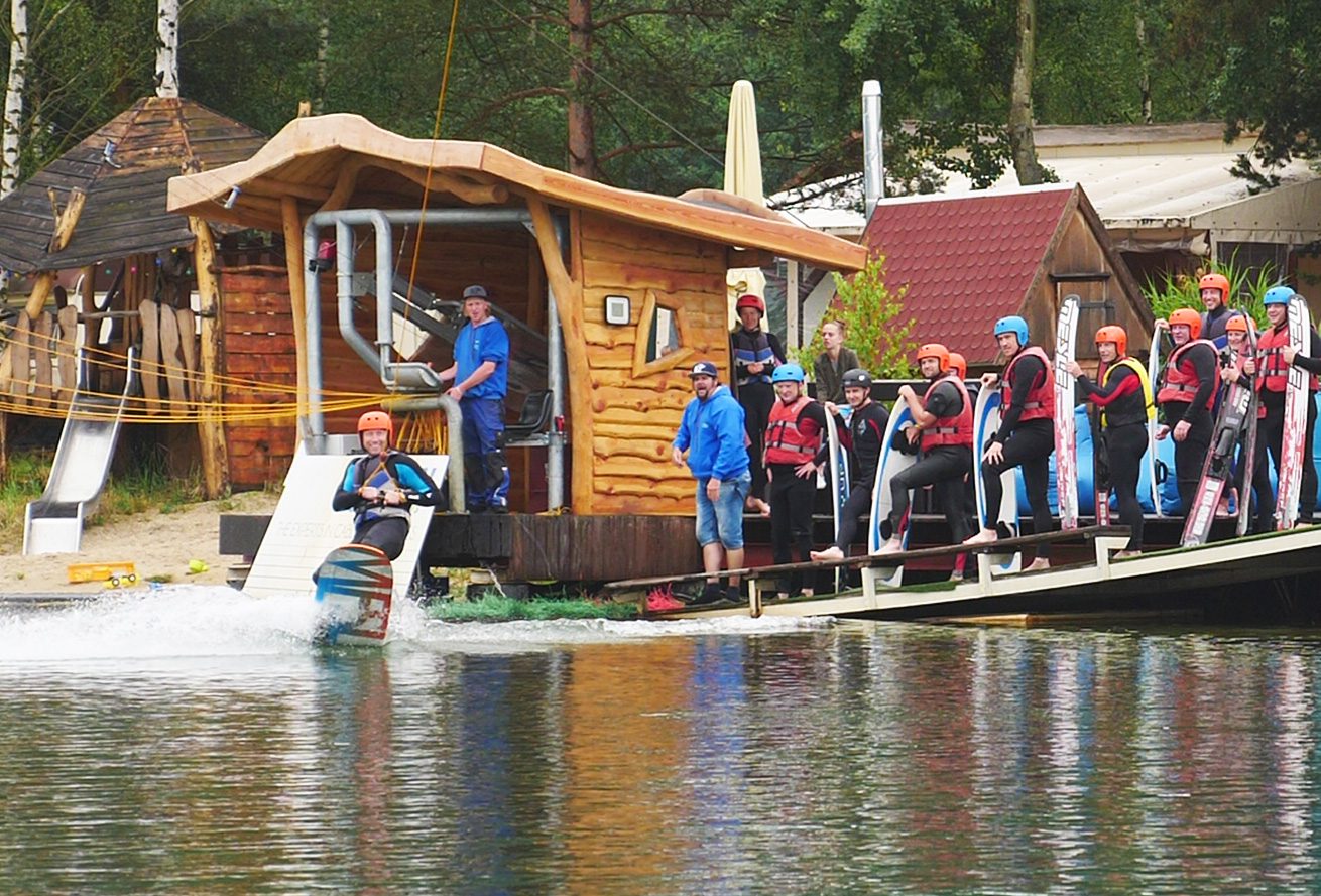 Gruppe mit Wasserski und Wakeboards schauen dem Anfänger beim fahren auf dem Wasser zu, während der Coach am Ufer noch Anweisungen gibt