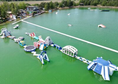 Riesiger Aquapark mit luftgepolsterten Elementen, die im Wasser stehen. Im Hintergrund ist die Wasserski- und Wakeboardanlage mit dem großen Außenbereich zu sehen