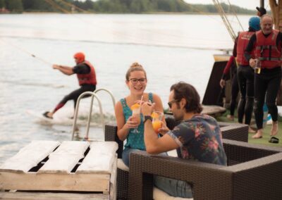 Frau und Mann sitzen am Wasser und trinken einen Cocktail, während im Hintergrund ein Wakeboarder im Wasser startet