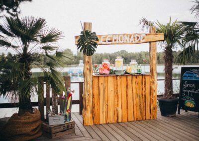 Aus Holz gebauter Lemonaden Stand mit liebevoll angerichteten Getränken neben Palmen und Blick auf den See