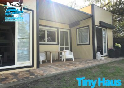 Tinyhaus mit offener Tür steht direkt am Strand, vor dem Haus stehen Sitzgelegenheiten