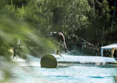 Junger Wakeboarder slidet entgegen der Fahrtrichtung auf einer dicken hohen Tube, die im Wasser steht